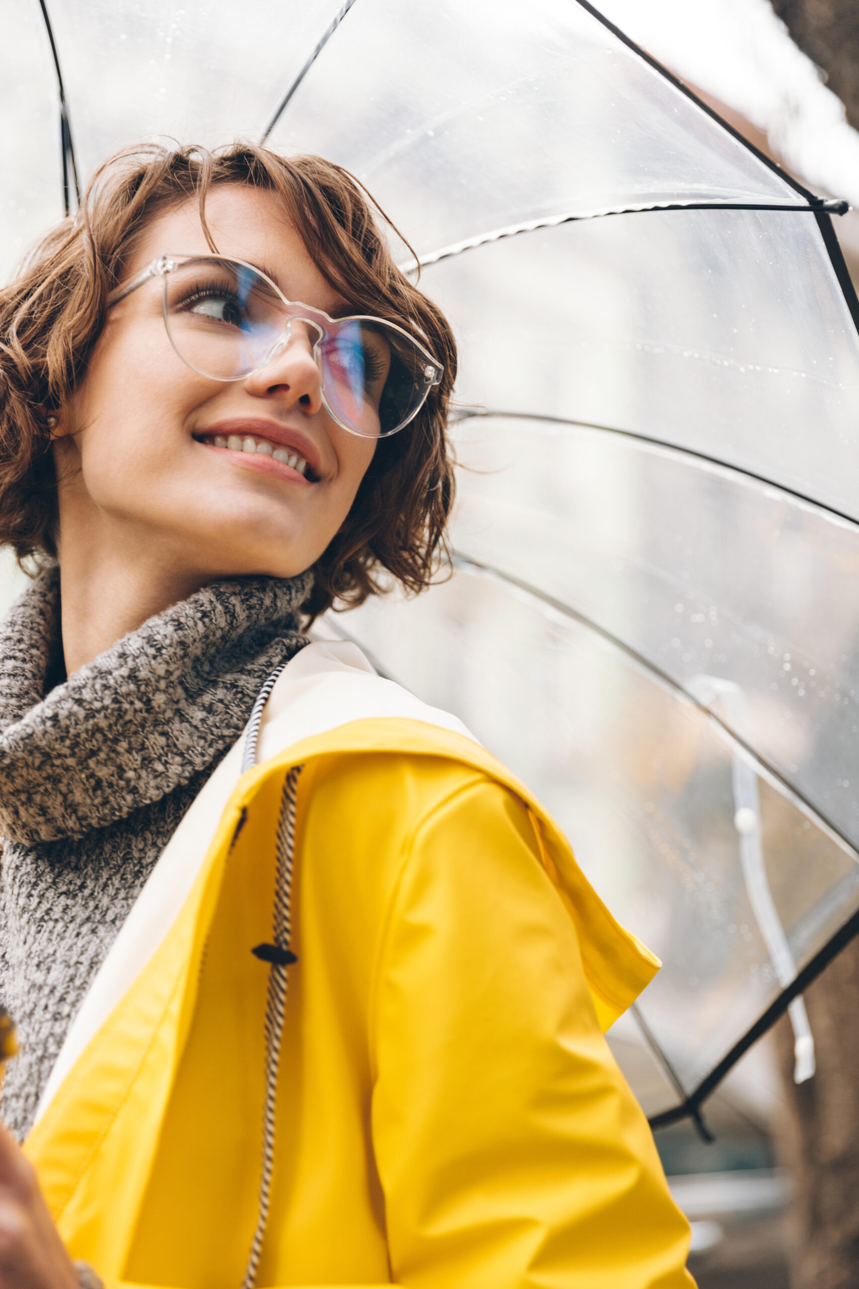 Smiling woman wearing clear eyeglasses and a yellow raincoat holding a transparent umbrella on a rainy day.
